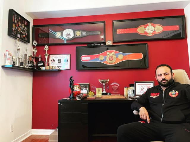 Roman Mitichyan seated in a red-walled room lined with championship belts and trophies