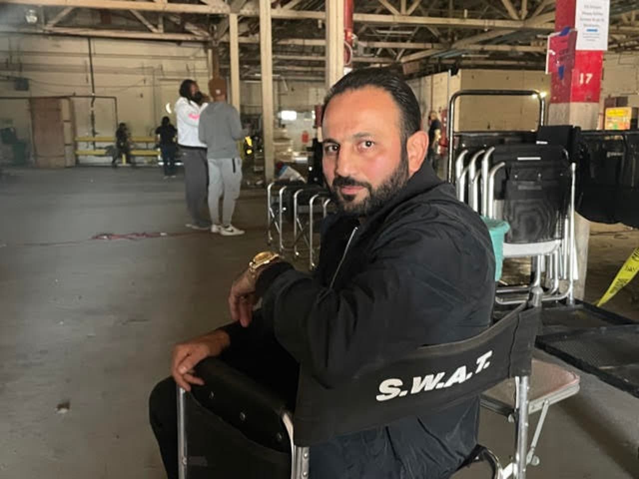 Roman Mitichyan seated in a chair marked S.W.A.T. on a warehouse set