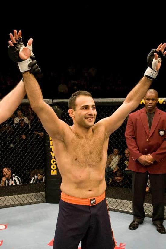 Roman Mitichyan raising both arms in victory inside the cage as the referee looks on